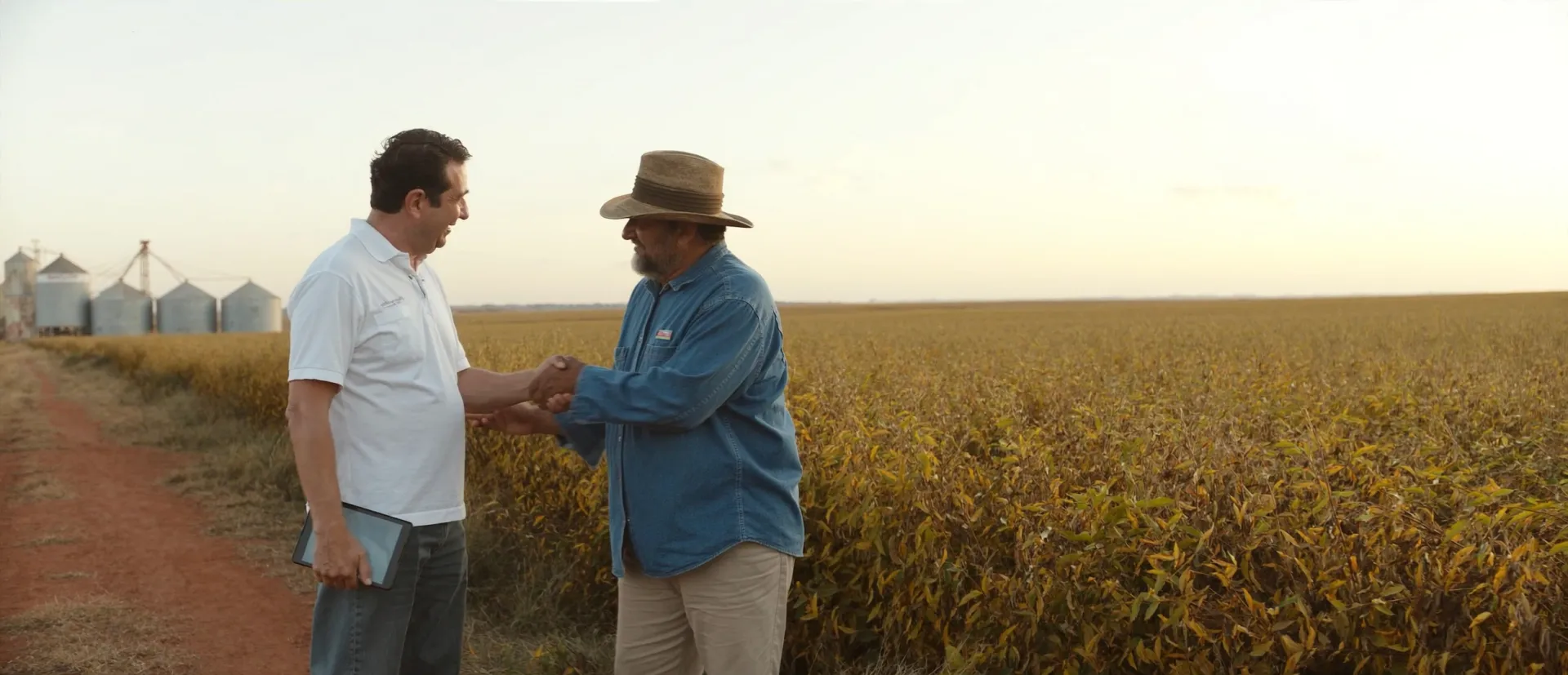 Un productor y un comprador se dan la mano junto a un campo de soja, con los silos al fondo